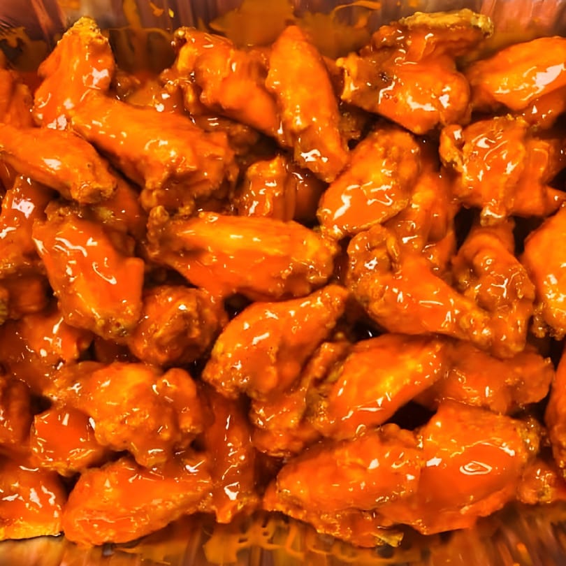 Close-up of a large batch of bright orange, saucy buffalo chicken wings in a foil pan.