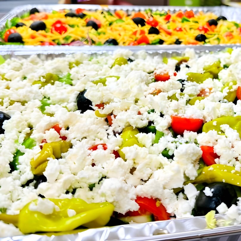 Two foil trays of salads, one with feta, peppers, tomatoes, and olives, the other with shredded cheese.